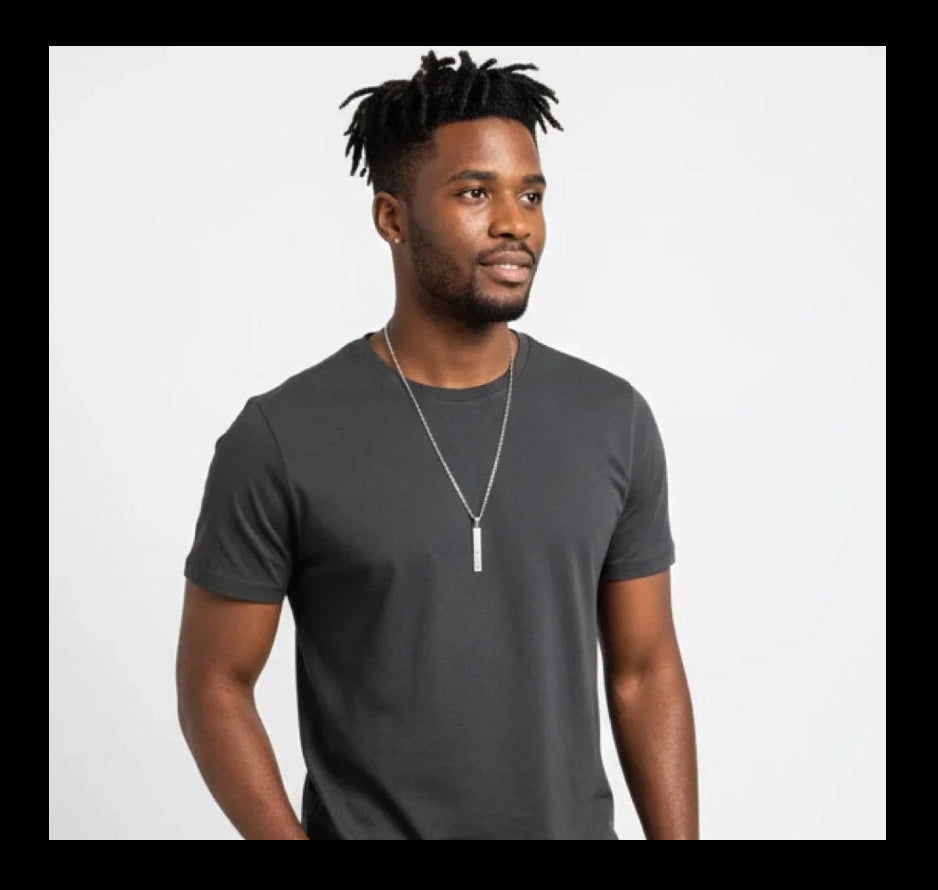 Man wearing dark gray t-shirt, slim blue jeans, white sneakers, and silver pendant necklace on white background