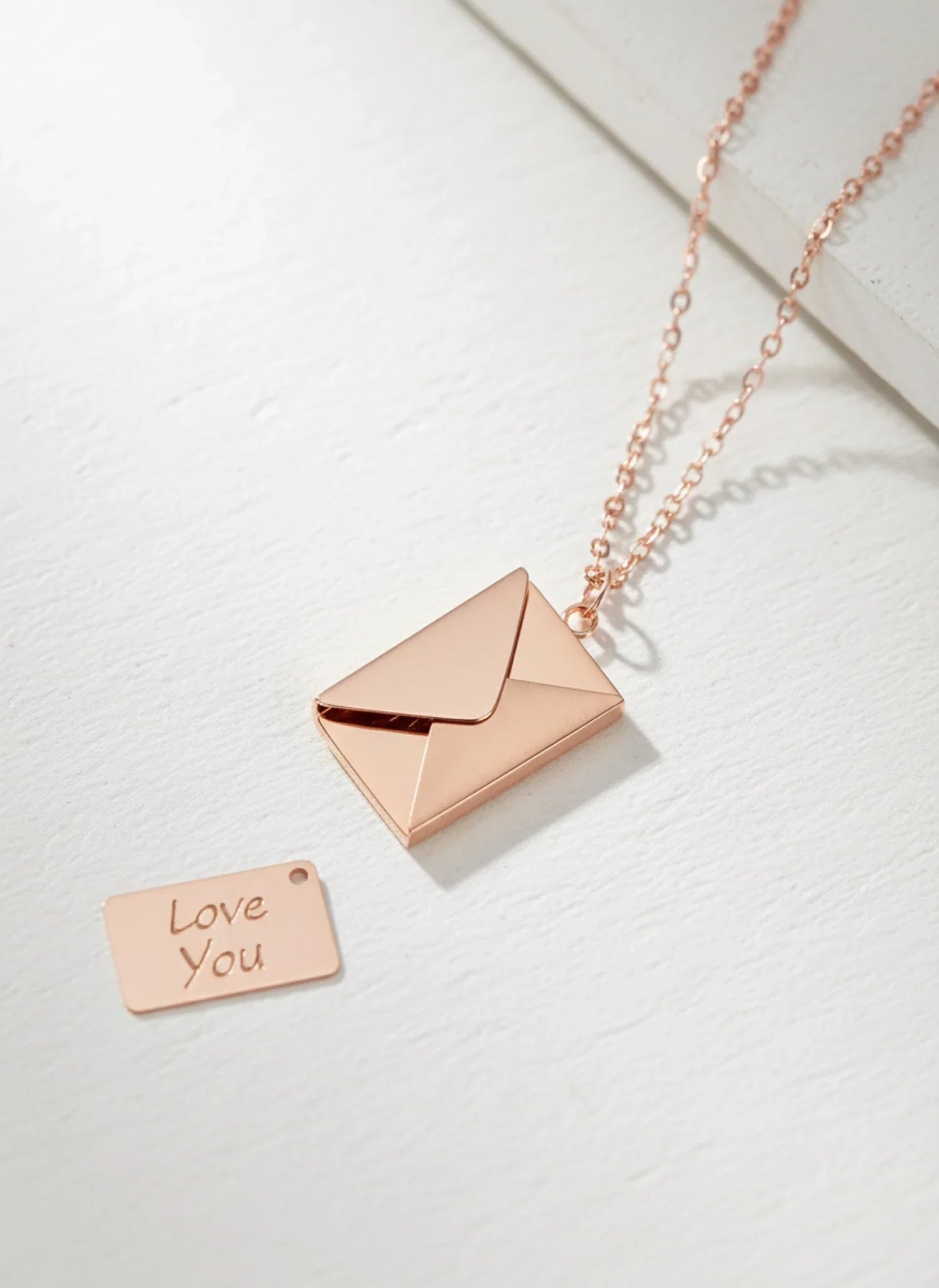 Rose gold envelope locket necklace with chain and small engraved tag reading 'Love You' on white surface